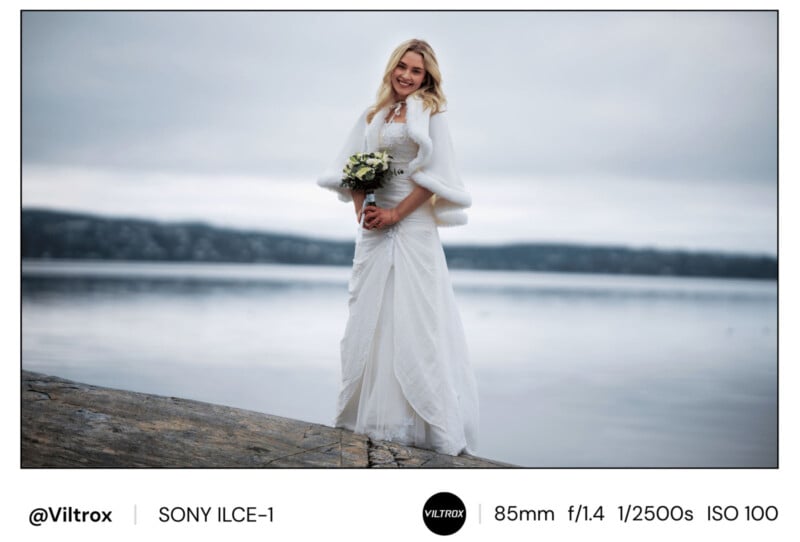A woman in a white wedding dress and shawl stands on a rocky shore holding a bouquet, smiling at the camera with a calm lake and distant hills in the background under an overcast sky.