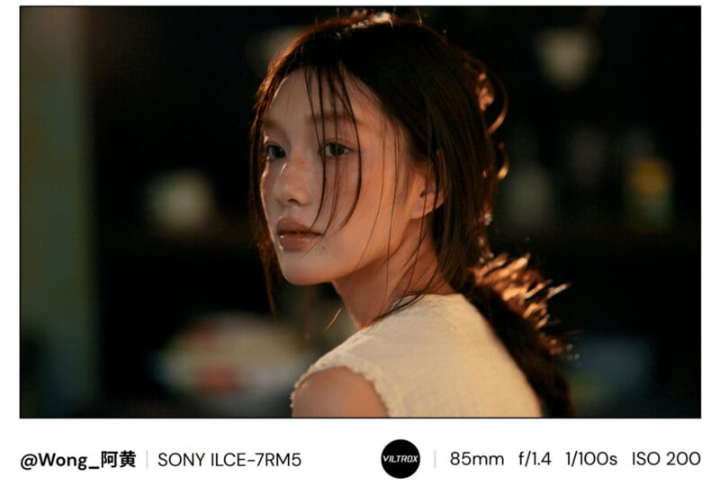 A young woman with long dark hair and light skin, wearing a white sleeveless top, looks over her shoulder in soft, warm lighting. The background is blurred. Camera details are labeled at the bottom of the image.
