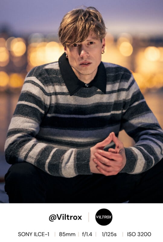 A young person with blonde hair, wearing a gray and black striped sweater and black collar, sits outdoors at dusk with blurred city lights in the background. Camera settings and Viltrox branding are shown below.