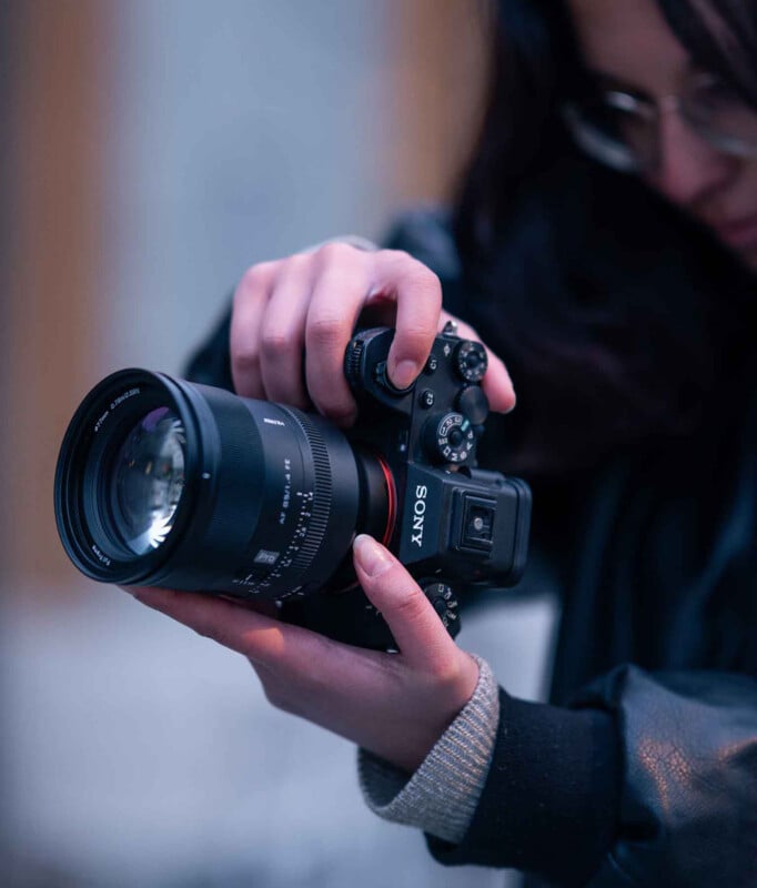 A person holding a Sony camera with both hands, adjusting the settings, with a blurred background and only part of their face visible.