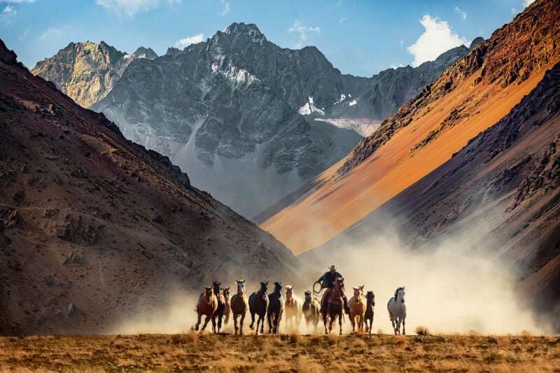 A person on horseback leads a group of running horses through a dusty valley, surrounded by steep, rugged mountains under a blue sky with scattered clouds.