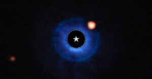 A bright orange planet appears in the upper right of the image, orbiting a central star obscured by a black circle, with faint blue rings of dust and gas surrounding the star against a dark background.