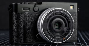 A close-up of a black Fujifilm camera with a textured grip and a Fujinon 23mm f/2.8 aspherical lens, resting on a grid-patterned surface against a dark background.