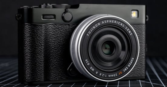 A close-up of a black Fujifilm camera with a textured grip and a Fujinon 23mm f/2.8 aspherical lens, resting on a grid-patterned surface against a dark background.