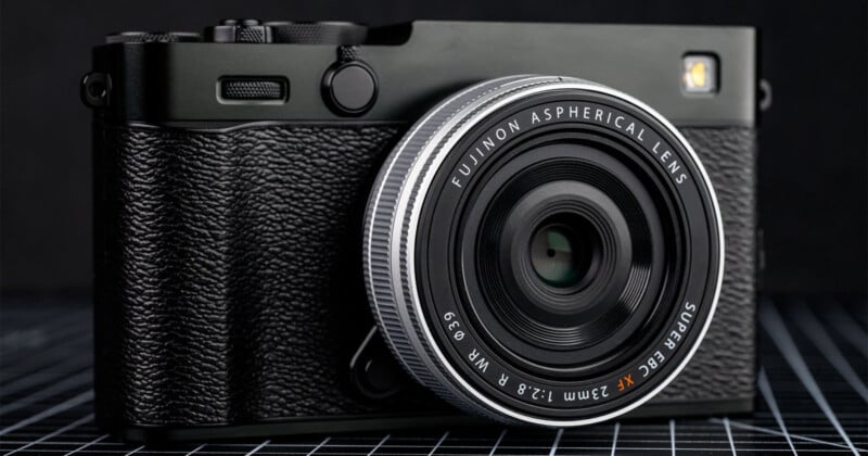 A close-up of a black Fujifilm camera with a textured grip and a Fujinon 23mm f/2.8 aspherical lens, resting on a grid-patterned surface against a dark background.