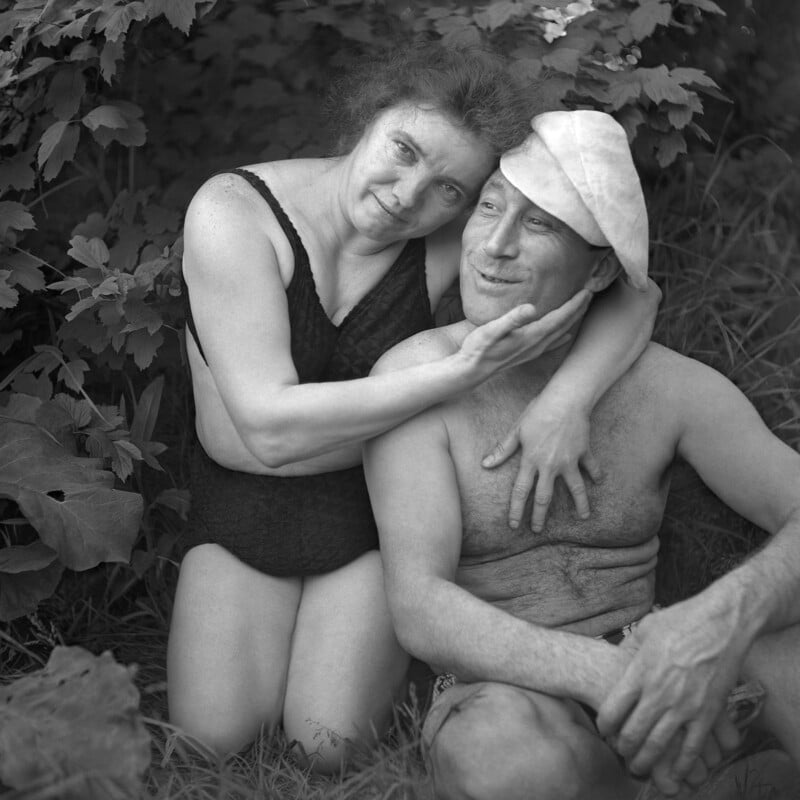 A woman in a black swimsuit embraces a smiling man wearing a white cap; they are sitting together outdoors among leafy plants and grass, both appearing relaxed and happy.