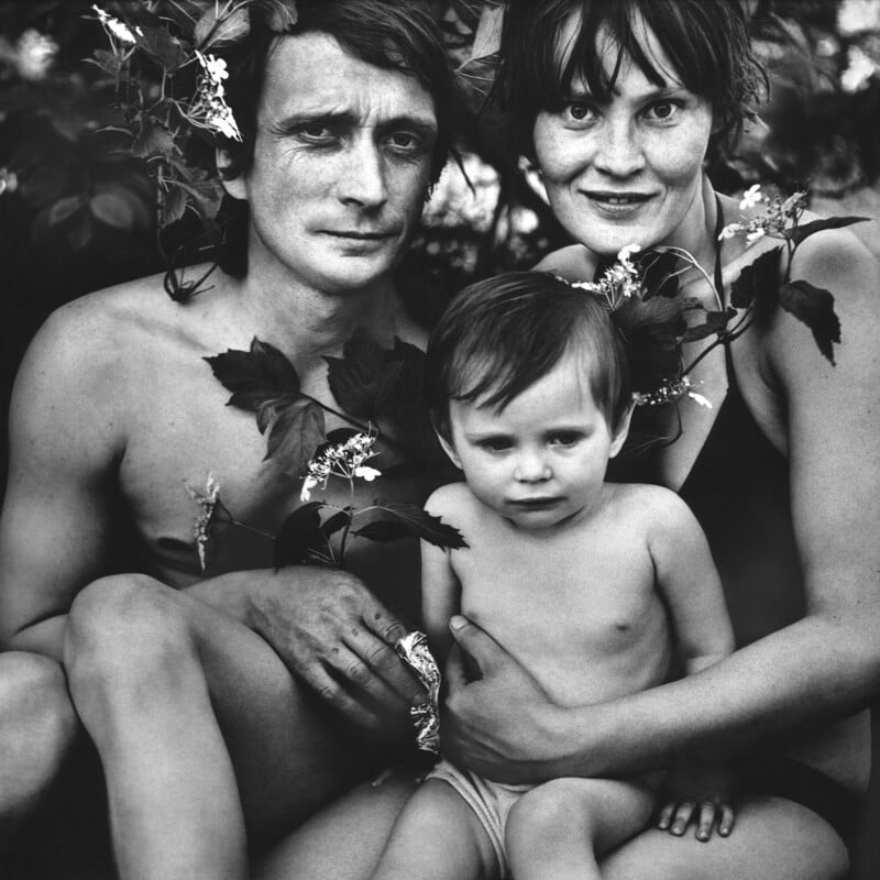 A black and white photo of two adults and a young child sitting closely together outdoors, surrounded by leafy branches and flowers. The adults are smiling gently and the child looks calmly at the camera.