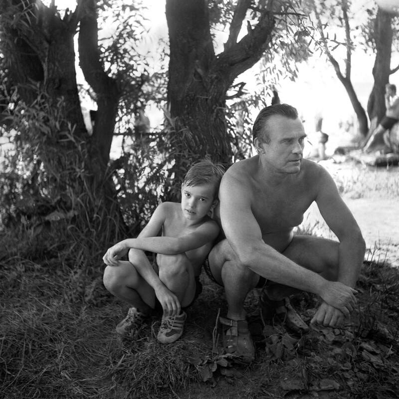 A black-and-white photo of a man and a young boy in swimsuits crouching by the base of a tree, looking thoughtfully into the distance, with sunlight filtering through the leaves.