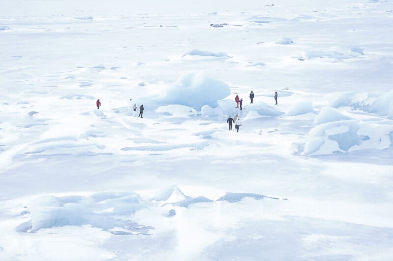 Several people walk and stand on a vast, snowy, and icy landscape surrounded by ice formations and patches of scattered snow under a bright, overcast sky.