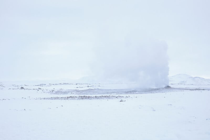 A snowy, white landscape with a cloud of steam or smoke rising from the ground, possibly from a hot spring or geyser, under an overcast sky. The scene appears cold and desolate.
