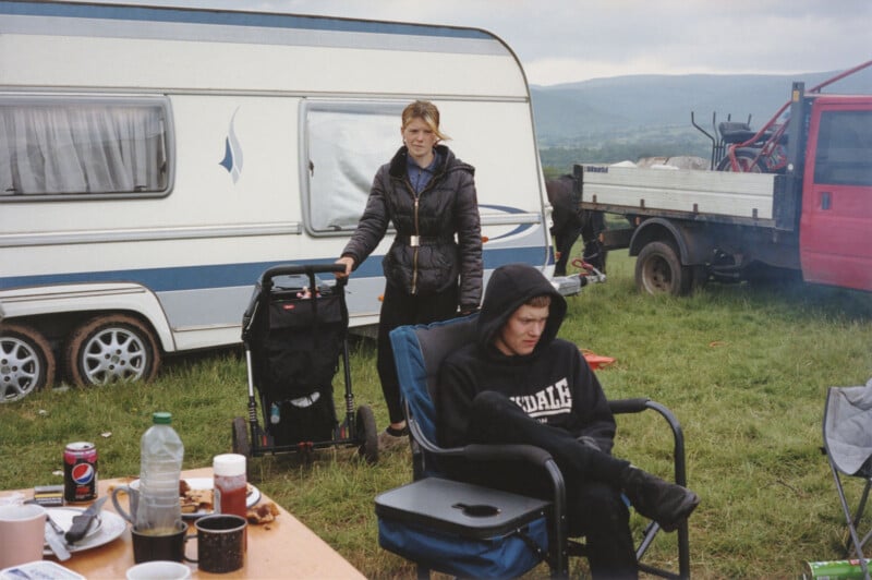 Two people are outdoors near a white caravan and a red truck. One stands with a stroller, wearing a jacket, while the other sits in a chair wearing a black hoodie. Food and drinks are on a table in the foreground.
