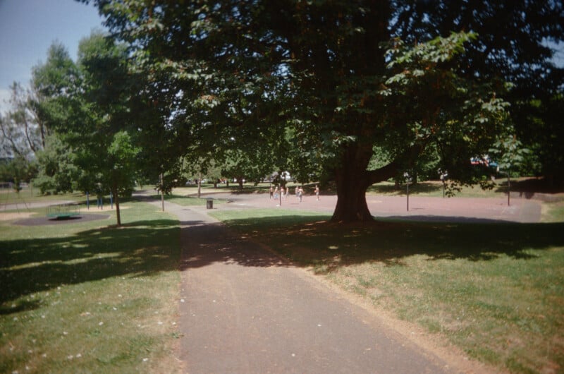 A paved path runs through a sunlit park with green grass, large leafy trees, and people in the distance. Shadows from the trees cover parts of the path, and playground equipment is visible in the background.