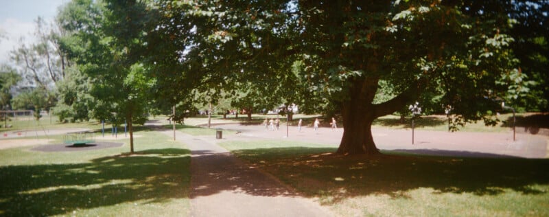 A sunny park scene with a paved path, large leafy trees, grassy areas, a playground to the left, and people walking or playing in the background under the shade.