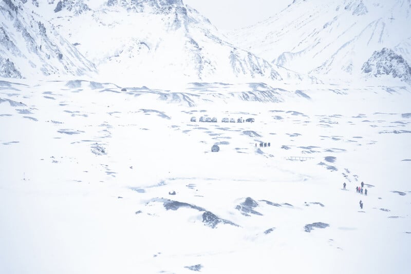 A snowy, mountainous landscape with scattered rocks and groups of people walking in the distance. The scene is dominated by white snow, with mountains rising in the background under an overcast sky.