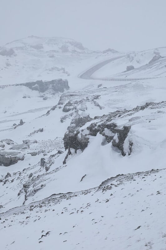 A snow-covered, rocky landscape with rolling hills and a winding road bordered by a fence, all shrouded in heavy snowfall and mist, creating a cold and desolate atmosphere.