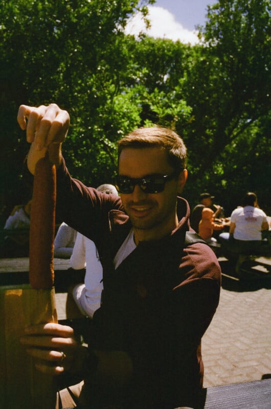A man wearing sunglasses and a dark jacket smiles while holding a long corn dog outdoors, with people sitting at picnic tables and green trees in the background.