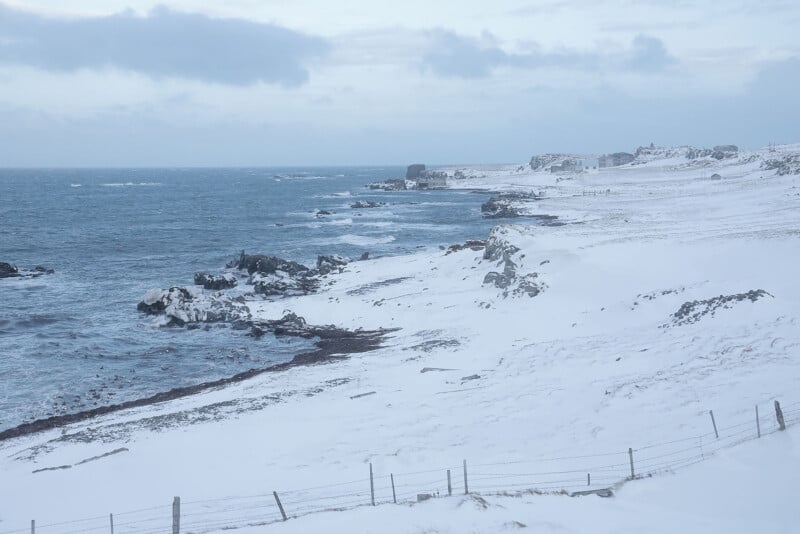 A snowy coastline with rocky outcrops meets the cold sea under a cloudy sky. A fence runs along the foreground, and the landscape appears windswept and desolate.