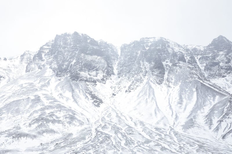 Snow-covered mountain peaks under an overcast sky, with rugged slopes and rocky ridges partially obscured by mist and snow. The scene appears cold and serene.