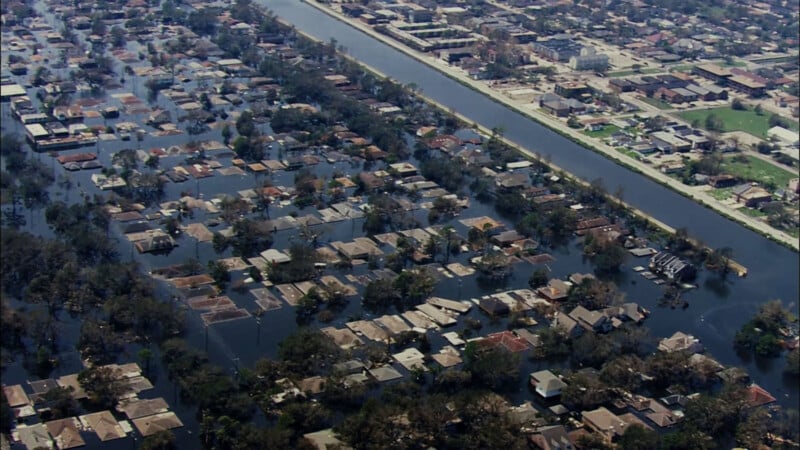 Aerial view of a flooded residential neighborhood, with water submerging streets and partially covering houses. A canal runs parallel to the area, and non-flooded buildings are visible in the upper part of the image.