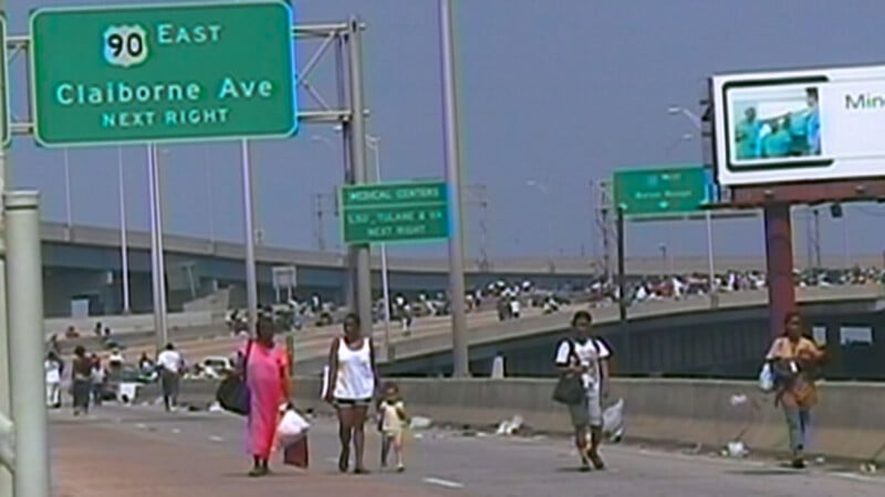 People carrying bags walk along a highway under green road signs for Claiborne Ave and Medical Center. A billboard and overpass crowded with more people are visible in the background. It is a sunny day.