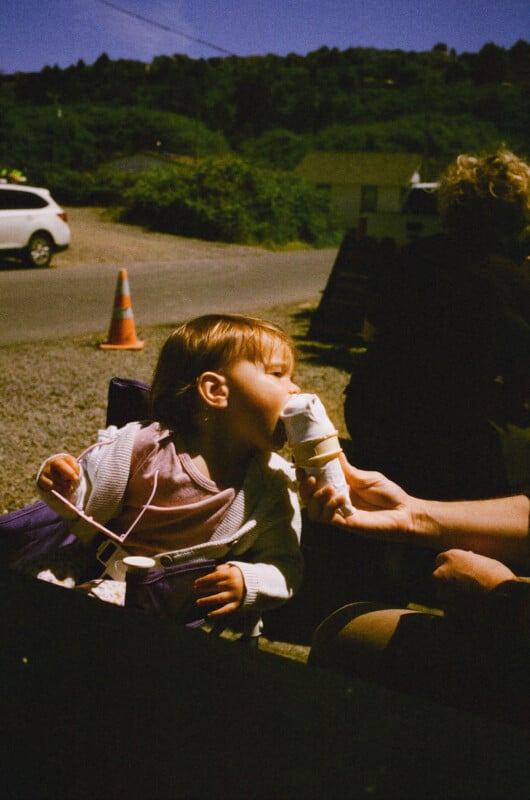 A young child sits outdoors and eagerly takes a bite of a vanilla ice cream cone held by an adult. A traffic cone and a white car are visible in the background.