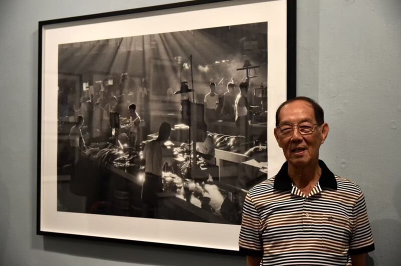 An elderly man in a striped shirt stands smiling next to a large, framed black-and-white photograph depicting a busy indoor scene with people working, lit dramatically by sunlight streaming through windows.