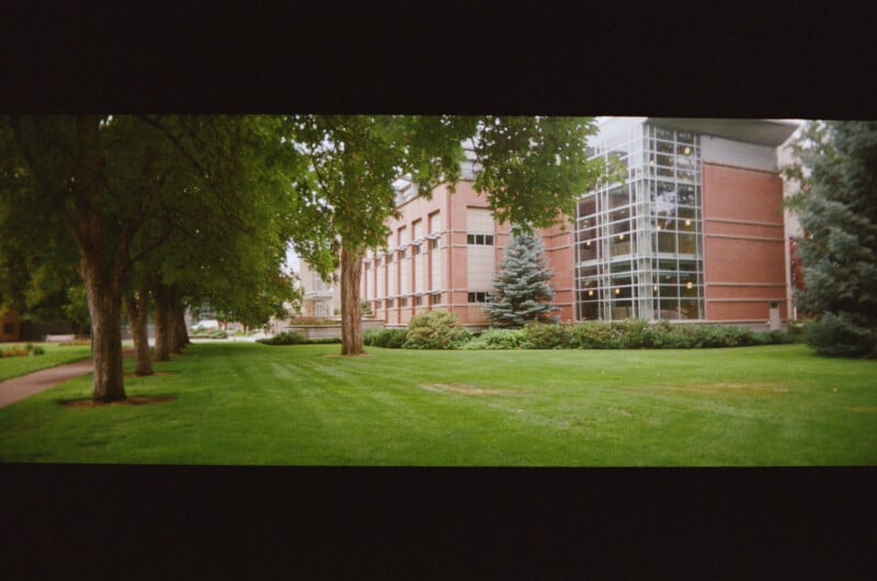 A modern brick building with large glass windows stands beside a green lawn and several trees; a sidewalk runs alongside the grass on a sunny day.