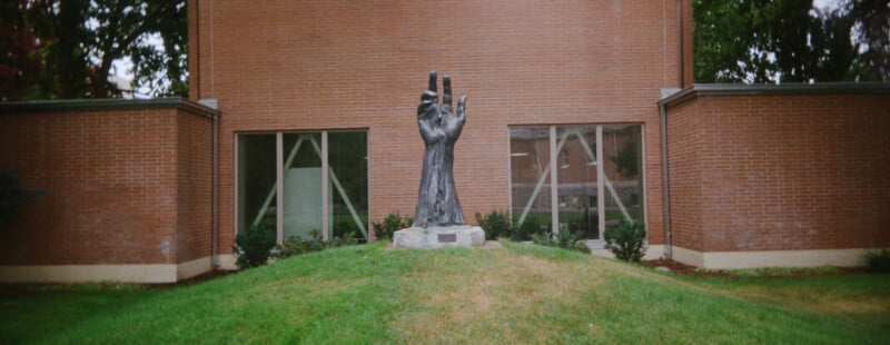 A large bronze sculpture of an upright hand stands on a grassy mound in front of a brick building with two windows and minimal landscaping.