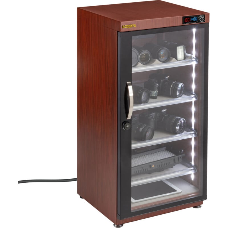A wooden electronic dry cabinet with a glass door displays cameras, lenses, and electronic devices on its shelves. The cabinet has an LED humidity display and is plugged into a power outlet.