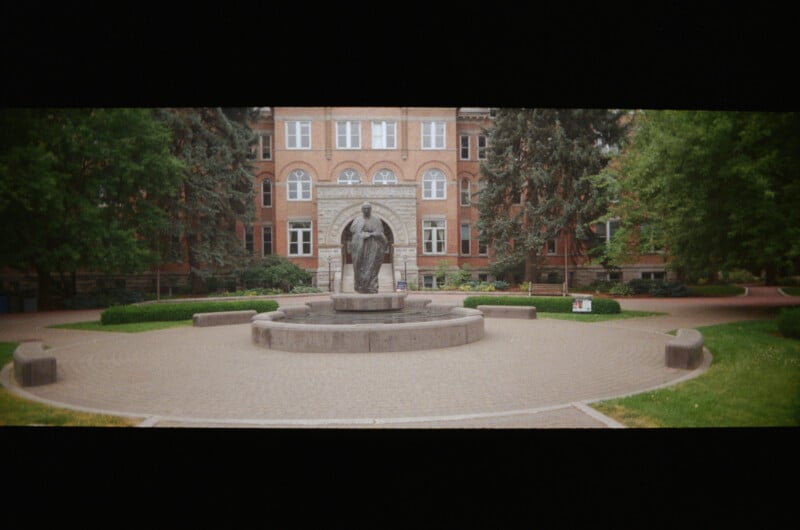 A statue stands on a circular stone pedestal in the center of a brick courtyard, surrounded by greenery, with a large brick building in the background.