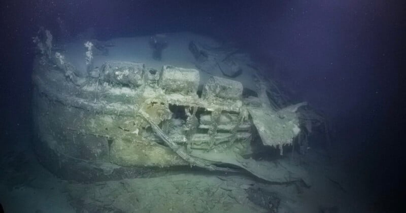 An underwater photo of a decaying shipwreck resting on the ocean floor, covered in sediment and marine growth, with parts of the hull and internal structure visible in murky blue water.