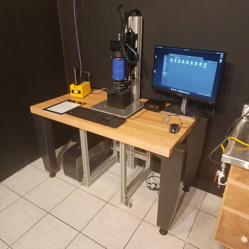 A desktop CNC machine with a blue spindle sits on a wooden table beside a computer monitor, keyboard, mouse, and a yellow control box in a tiled room with black walls.