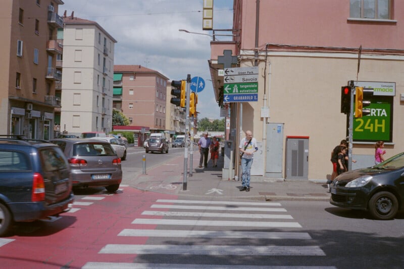 A city street intersection with cars stopped at a crosswalk, pedestrians walking and waiting, street signs, traffic lights, and buildings, including a store with a green "APERTA 24H" sign.