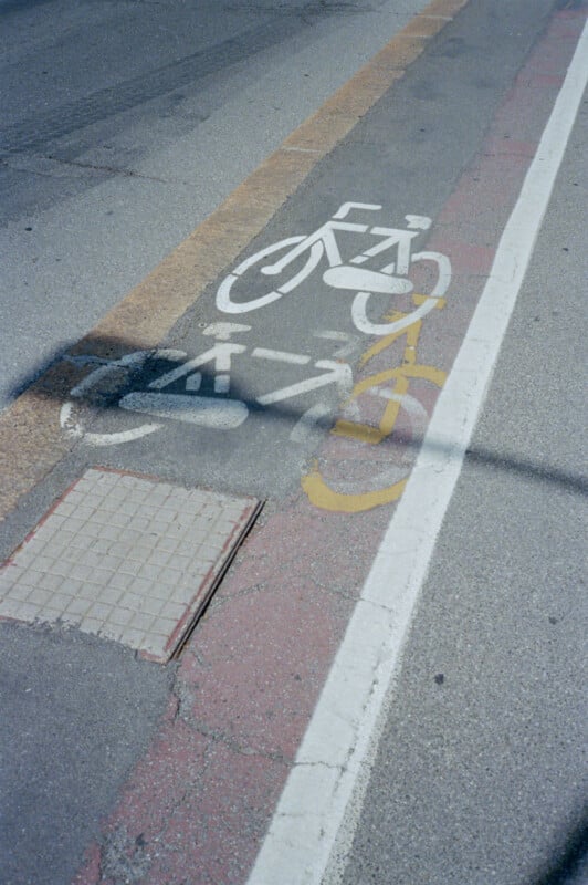 A white bicycle lane symbol on a city street is partially covered by the shadow of a bicycle-shaped sign, creating overlapping bike silhouettes on the pavement. A tactile paving tile is nearby.