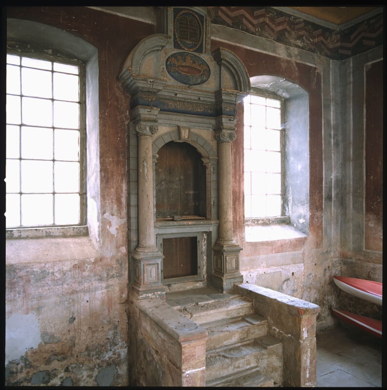 A weathered, ornate wooden pulpit with steps stands between two large, sunlit windows in an old, faded church interior with peeling paint and worn walls. A red bench is partially visible on the right.