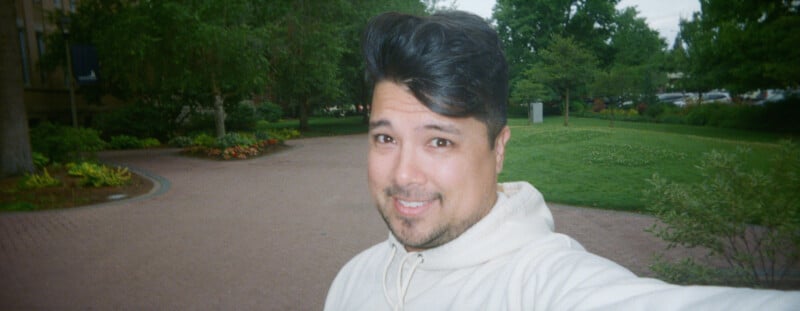 A man with dark hair and a short beard, wearing a beige hoodie, takes a selfie outdoors in a green park with trees, grass, and flower beds in the background.