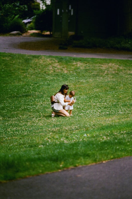 A woman kneels on a grassy field beside a young child. Both are looking down, possibly at something on the ground, surrounded by green grass with a path and trees in the background.
