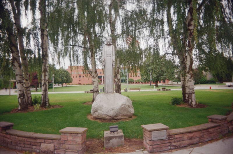 A stone statue of a figure stands on a large rock, surrounded by birch trees and a low stone wall in a grassy park, with a red brick building visible in the background.
