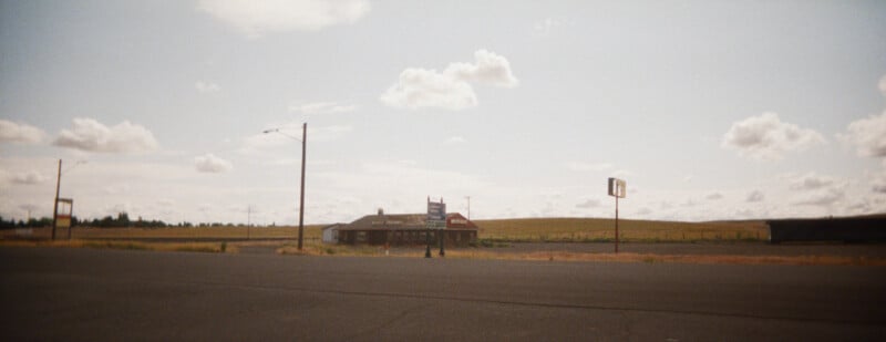A small, isolated building and empty signs stand beside a wide, deserted road under a partly cloudy sky, with open fields and low hills in the background.