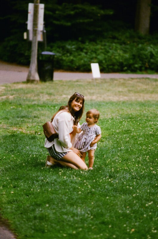 A woman kneels on grass smiling and holding hands with a young child in a striped outfit in a park, with trees and a walking path in the background.