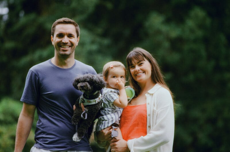 A smiling man, woman, and small child stand together outdoors with a small black dog in the man's arms. The child has a finger in their mouth. Lush green foliage is blurred in the background.