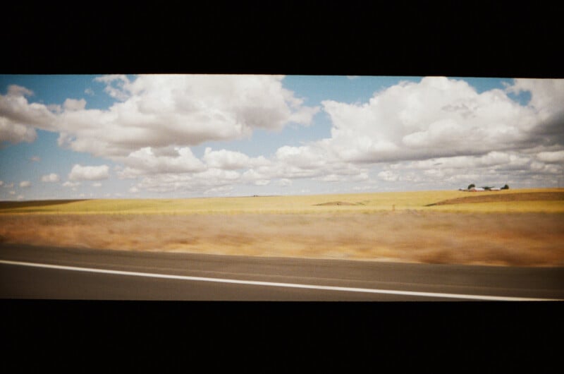 Wide view of an open landscape seen from a moving vehicle, with a blurred highway in the foreground, golden fields in the middle, and a sky full of large white clouds.