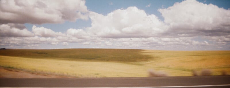 A scenic view of rolling grassy fields under a partly cloudy blue sky, seen from the roadside. The landscape appears sunlit with soft, fluffy clouds scattered above.