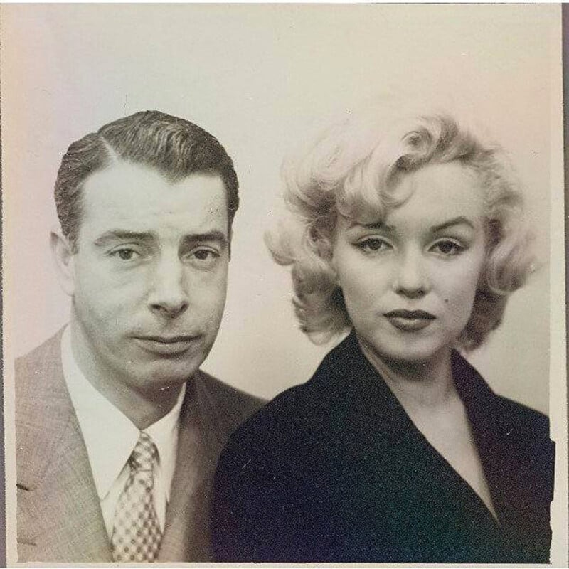 A black-and-white portrait of a man in a suit and tie next to a woman with short, wavy blonde hair and a dark outfit, both looking directly at the camera with neutral expressions.