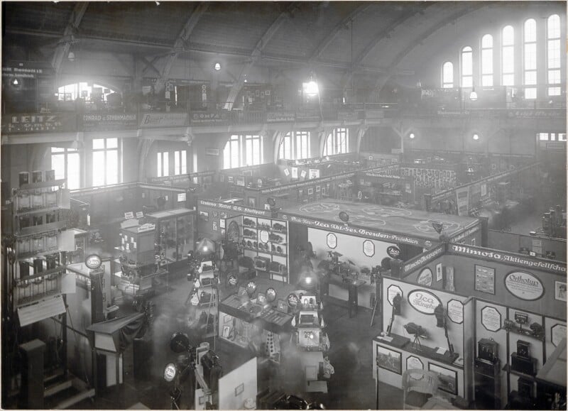 Wide-angle view of a large, early 20th-century exhibition hall with high arched ceilings, sunlight streaming through tall windows, and numerous booths displaying various equipment and products.