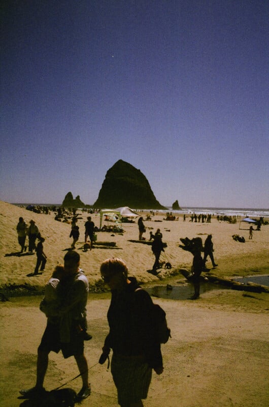People walk along a beach with a large rock formation in the background under a clear blue sky. The beach is crowded with visitors, some sitting under umbrellas, and the sun casts strong shadows.