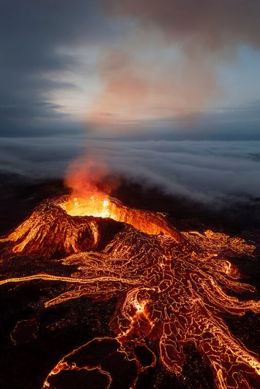Aerial view of an erupting volcano at dusk, with glowing lava flows streaming down its sides and thick smoke rising into a cloudy sky.