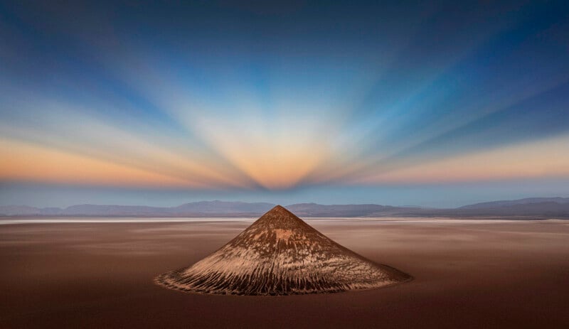A lone conical volcano rises from an expansive desert plain, with dramatic sun rays fanning out across the sky at sunrise or sunset, casting soft pastel colors above distant mountains.