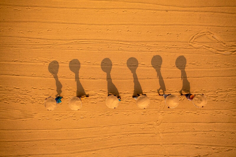 Aerial view of six people wearing conical hats walking in a line across golden sand, casting long, distinct shadows behind them; footprints and tracks are visible on the sand.