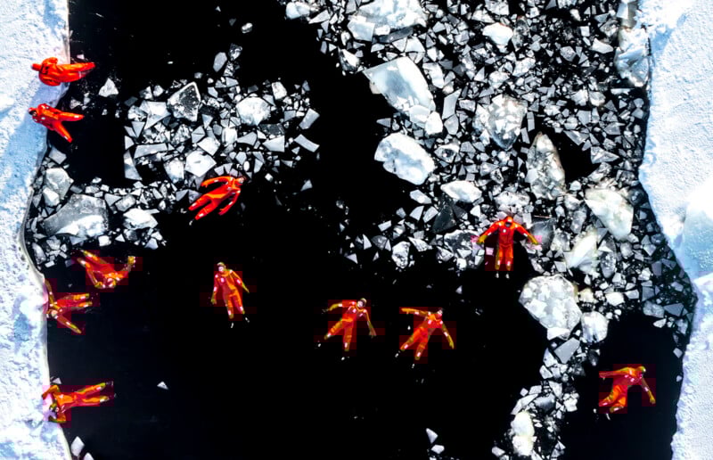 Aerial view of people in bright red suits floating on dark, icy water surrounded by large chunks of white ice, with snowy ground along the edges.